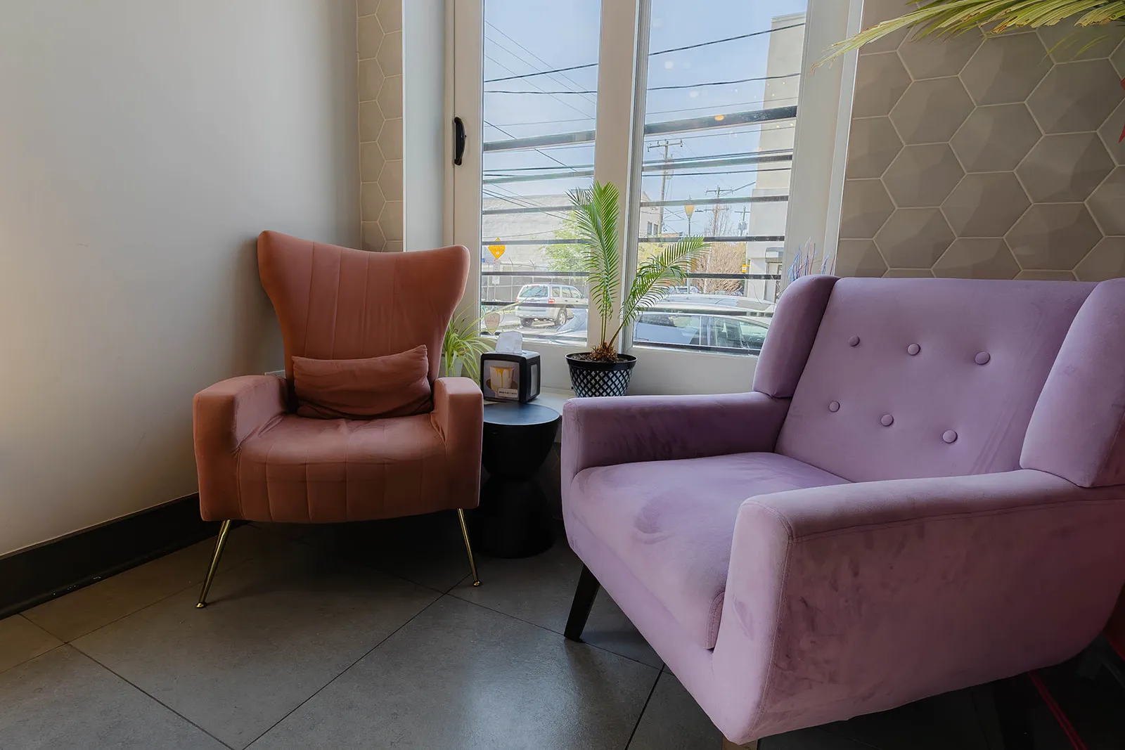 A pink wingback and a lilac loveseat in the window, with a palm between them